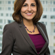 Neera Tanden of the Center for American Progress smiles and stands in front of a window wearing a grey blazer, dark purple top and a necklace.